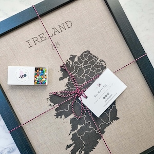 Może przedstawiać: Oprawione dzieło sztuki przedstawiające mapę Irlandii w ciemnoszarym kolorze, z napisem „IRELAND” i rokiem „2024”. Dołączone jest małe pudełko z kolorowymi szpilkami i etykieta z logo pszczoły. Rama jest czarna.