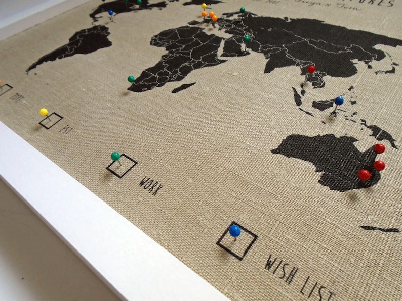 May include: A world map with push pins marking locations. The map is on a beige linen background with a white frame. The map has text that says "Wish List" and "Work".