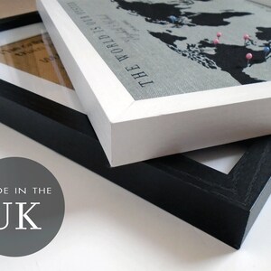 May include: Two picture frames, one black and one white, are stacked on top of each other. The white frame has a world map print with the text "The World Is Our Home" and pink and blue push pins. The black frame has a blank white background. The text "Made in the UK" is printed on a gray circle in the lower left corner of the image.
