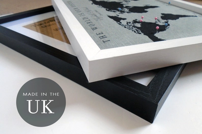 May include: Two picture frames, one black and one white, are stacked on top of each other. The white frame has a world map print with the text "The World Is Our Home" and pink and blue push pins. The black frame has a blank white background. The text "Made in the UK" is printed on a gray circle in the lower left corner of the image.