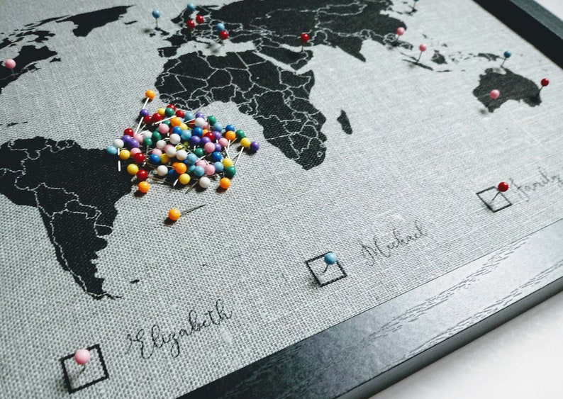 May include: A world map with a black and white design, framed in black wood. The map is covered in colorful push pins, with a large cluster of pins in the center of the map. The map has text that says "Elizabeth", "Michael", and "Jennifer".