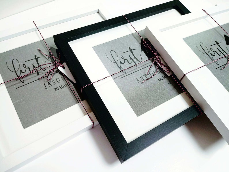 May include: Three white picture frames with black mounts, each containing a print with the word "first" and a name. The frames are tied together with red and white twine.