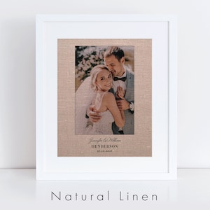May include: A framed wedding photograph printed on natural linen with a white frame. The photo shows a couple embracing, with the bride wearing a white dress and the groom wearing a suit. The text below the photo reads "Jennifer & William HENDERSON 07.18.2012".