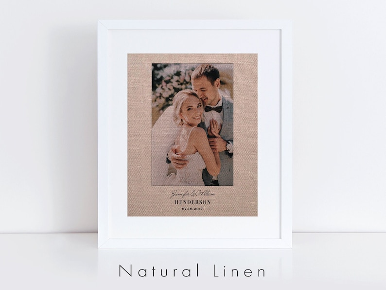 May include: A framed wedding photograph printed on natural linen with a white frame. The photo shows a couple embracing, with the bride wearing a white dress and the groom wearing a suit. The text below the photo reads "Jennifer & William HENDERSON 07.18.2012".