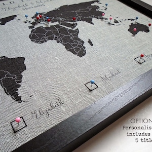 May include: A black and white world map printed on a gray linen fabric with push pins in various colors. The map is framed in black wood. The text "WORLD" is at the top of the map. The text "Elizabeth & 29.01.2005" is below the word "WORLD". The text "Elizabeth" is written in cursive at the bottom of the map. The text "Michael" is written in cursive in the upper right corner of the map. The text "family" is written in cursive in the upper right corner of the map. The text "OPTIONAL: Personalised key includes upto 5 titles" is written in black text at the bottom right corner of the image.
