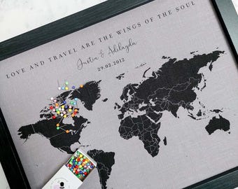 May include: A framed world map with a grey linen background and black outlines of countries. The map is covered in colourful push pins and the text "Love and travel are the wings of the soul" is printed at the top. The text "Justin & Adelayda 29.02.2012" is printed below the map.