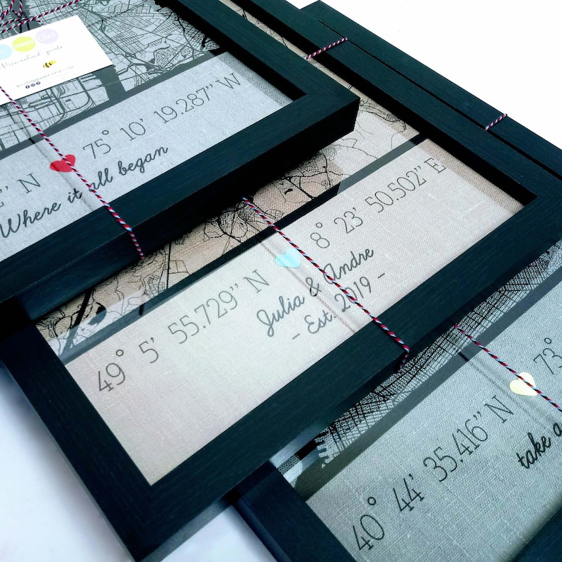 May include: Three framed maps with black frames and white string. Each map has a different location with coordinates and a personalised message. The first map says "Where it all began" with coordinates 75&deg; 10' 19.287" W. The second map says "Julia & Andre - Est. 2019 -" with coordinates 49&deg; 5' 55.729" N and 8&deg; 23' 50.502" E. The third map says "take a" with coordinates 40&deg; 44' 35.416" N.