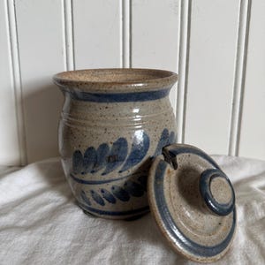 May include: A handmade ceramic jar with a lid. The jar is beige with blue floral designs and a blue rim. The lid is also beige with a blue rim and a small handle. The jar is sitting on a white cloth.