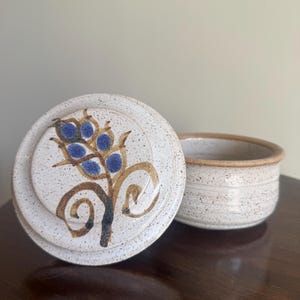 May include: A ceramic butter dish with a lid featuring a floral design in blue and brown. The dish and lid are speckled with a neutral color palette. The lid is off to the side, revealing the interior of the dish.