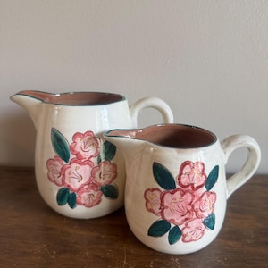 May include: Two ceramic pitchers with a cream-colored base and a floral design. The flowers are pink with green leaves. The rims and interiors are brown. The pitchers have handles and spouts.