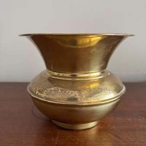 May include: A polished brass bowl with a wide, flared rim and a rounded, bulbous body. The metal surface displays a warm, metallic gold tone with visible signs of natural oxidation and patina, particularly around the midsection of the vessel.