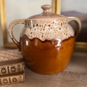 May include: A vintage ceramic pot with a lid, featuring a two-tone glaze in shades of brown and tan. The pot has two handles and a textured, lace-like pattern around the rim. The pot is sitting on a wooden surface.