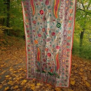 Puede incluir: Un textil plateado con intrincados bordados, con un diseño floral y paisley en rojo, verde, naranja y amarillo. Los bordes están ribeteados con un borde rojo. El textil se exhibe al aire libre, colgado contra un telón de fondo de árboles.