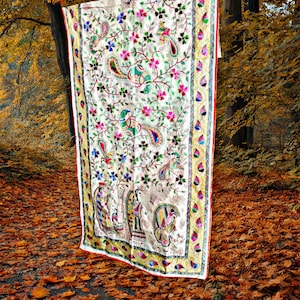 Puede incluir: Un textil color crema con intrincados bordados con diseños de cachemir, florales y de pavo real en colores vibrantes. El borde tiene un ribete dorado y rojo. El textil está colgado al aire libre, rodeado de follaje otoñal.