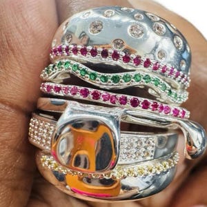 Peut inclure: Une bague argentée originale en forme de hamburger. Le pain supérieur est orné de pierres transparentes, tandis que les couches présentent des pierres précieuses rouges et vertes. Le pain inférieur est bordé de pierres jaunes, créant un bijou amusant.