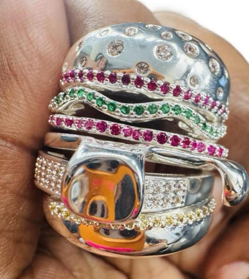 Pu&ograve; includere: Un anello d'argento unico progettato per assomigliare a un hamburger. La parte superiore del panino &egrave; tempestata di pietre trasparenti, mentre gli strati includono pietre preziose rosse e verdi. La parte inferiore del panino &egrave; bordata con pietre gialle.