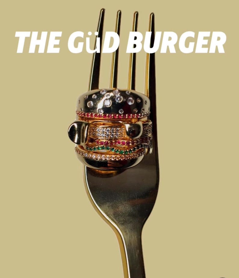 May include: A gold-colored fork holding a burger-shaped ring made of multiple rings. The burger ring has a top bun with clear stones, a red stone layer, and a layer with clear and green stones. The text "THE G&Uuml;D BURGER" is at the top.