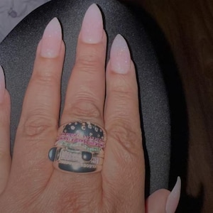 Pu&ograve; includere: Un anello d'argento a forma di hamburger, con la parte superiore nera, strati rosa e verdi e pietre trasparenti. L'anello &egrave; su una mano con unghie lunghe rosa chiaro.