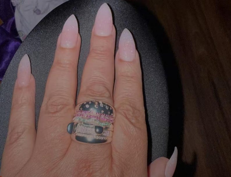 Pu&ograve; includere: Un anello d'argento a forma di hamburger, con la parte superiore nera, strati rosa e verdi e pietre trasparenti. L'anello &egrave; su una mano con unghie lunghe rosa chiaro.