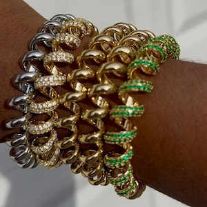 Op de afbeelding: Een verzameling spiraalvormige armbanden in goud, zilver en goud met groene of heldere edelstenen. De armbanden zijn op een pols gestapeld en tonen hun unieke ontwerp en verschillende kleuren.