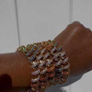 May include: A set of gold-toned metal bracelets featuring a spiral design. Each bracelet is encrusted with small, multi-coloured gemstones in shades of blue, green, orange, and clear, creating a vibrant, sparkling appearance across the wrist.