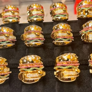 May include: A collection of gold-toned rings designed to resemble miniature hamburgers. Each ring features a bun, patty, and colorful gemstone accents, including clear, pink, and green stones. The rings are displayed on a black surface.