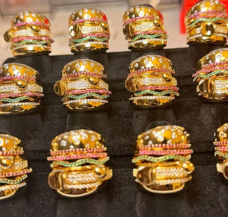 May include: A collection of gold-toned rings designed to resemble miniature hamburgers. Each ring features a bun, patty, and colorful gemstone accents, including clear, pink, and green stones. The rings are displayed on a black surface.