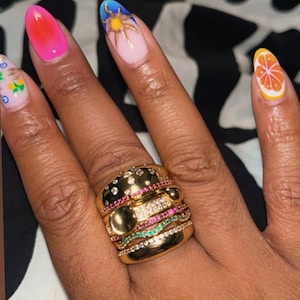 May include: A hand with long, oval-shaped nails, each painted with a different design. A gold ring with multiple bands and gemstones is on the ring finger. The nails feature a gradient, a sun, and a citrus fruit.