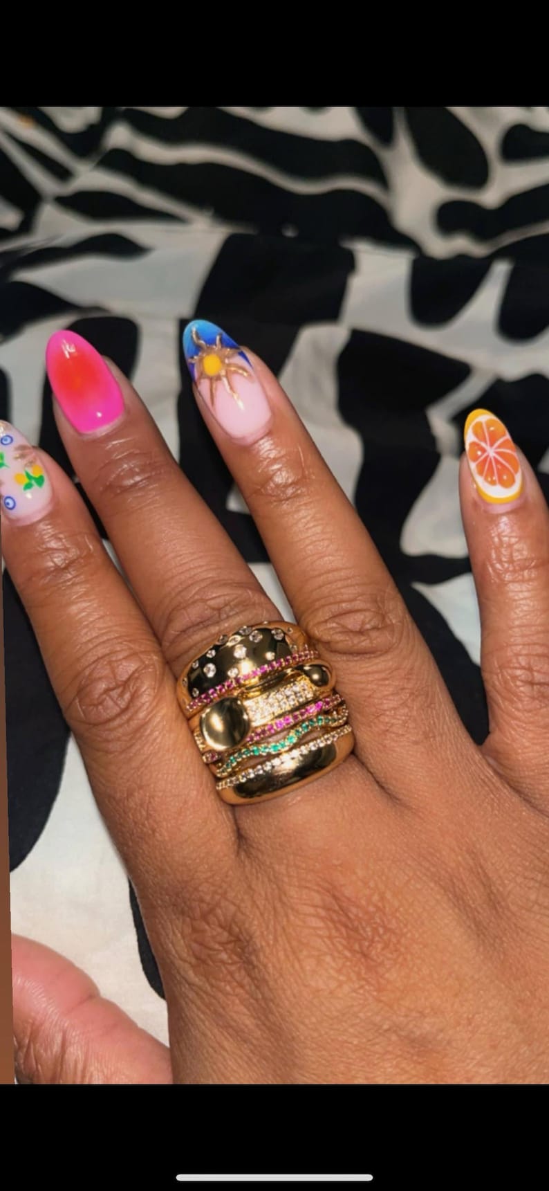 May include: A hand with long, oval-shaped nails, each painted with a different design. A gold ring with multiple bands and gemstones is on the ring finger. The nails feature a gradient, a sun, and a citrus fruit.