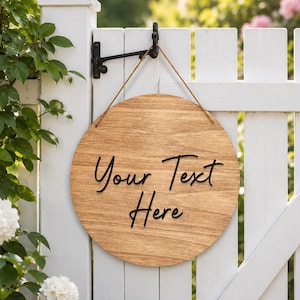 Puede incluir: Un letrero circular de madera con un acabado natural de veta de madera marrón claro, que presenta el texto negro en relieve "Your Text Here" en letra cursiva. El letrero está suspendido por un cordel marrón fino de un soporte de gancho de metal negro.