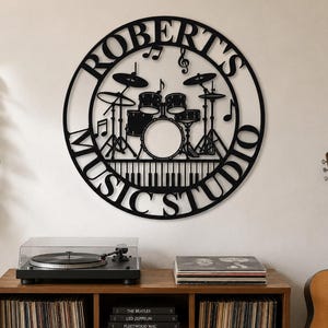 Puede incluir: Un letrero de pared circular de metal negro con la silueta de una batería, teclas de piano en la parte inferior y notas musicales. El texto "ROBERT'S MUSIC STUDIO" está integrado en el marco circular que rodea el diseño central.