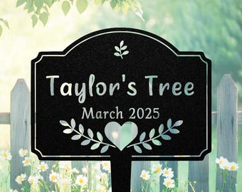 May include: Black metal garden stake with the text "Taylor's Tree March 2025". The sign has a decorative border and a heart-shaped cutout. The background includes a wooden fence and flowers.