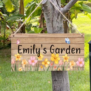 Pode incluir: Placa de madeira com o texto "Emily's Garden" em letras pretas. A placa apresenta uma borda floral com flores amarelas, rosas e laranjas e duas borboletas. A placa está pendurada em uma árvore.
