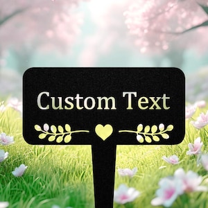 May include: A black metal garden stake with the words "Custom Text" in a light colour. The stake features a yellow heart and leaf accents. The background shows a blurred garden scene with flowers.
