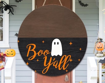 May include: Round wooden Halloween sign with a brown top and black bottom. The black section features a white ghost and the orange text "Boo Y'all". The sign is hanging on a front porch decorated for Halloween.