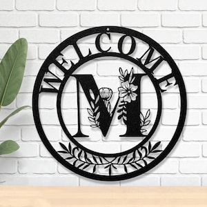 May include: Black metal welcome sign with the letter "M" in the center, adorned with floral and leaf details. The word "WELCOME" is cut out around the outer circle. The sign is designed to be hung on a wall.