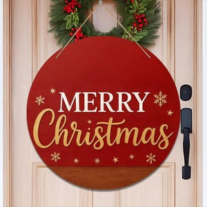 May include: A round, red wooden Christmas sign with the words "MERRY Christmas" in white and gold script. The sign is decorated with snowflakes and stars. It hangs on a door with a green wreath in the background.