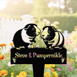 May include: Black metal garden stake featuring two guinea pig silhouettes. The stake has the names "Steve & Pumpernikle" in colorful text, with paw prints on either side. The background includes a white picket fence and colorful flowers.