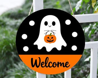 May include: Circular Halloween welcome sign with a black upper section and an orange lower section. A white ghost holding an orange pumpkin is in the center. The word "Welcome" is written in black on the orange section. White dots surround the ghost.
