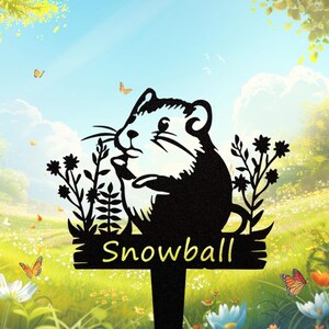 Könnte beinhalten: Schwarzer Metall-Gartenstecker mit einem Murmeltier-Design und dem Namen "Snowball". Das Murmeltier hält Blumen. Der Stecker steht in einem Garten mit Schmetterlingen und Blumen unter blauem Himmel.
