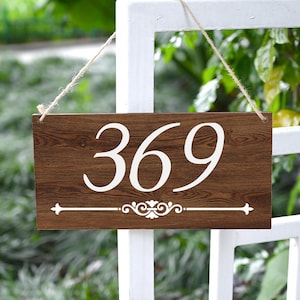 May include: A rectangular wooden sign with the numbers "369" in white script. The sign has a dark brown wood grain finish and a white decorative flourish at the bottom. It hangs from a white fence with a natural twine.