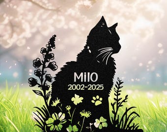 May include: Black metal cat silhouette garden stake with the name "Milo" and the years 2002-2025. The cat is surrounded by floral and plant cutouts, and a paw print. The background shows green grass and a blurred, flowering tree.