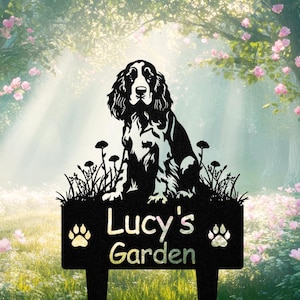 Pode incluir: Placa de jardim em metal preto com a silhueta de um cão sentado sobre a relva com flores. A placa diz "Lucy's Garden" em escrita branca, com pegadas de patas. O fundo mostra um jardim com flores cor-de-rosa.