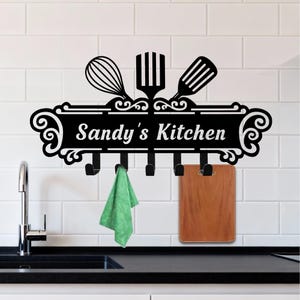 May include: Black metal kitchen wall decor with the text "Sandy's Kitchen" in white. The decor features a whisk, spatula, and slotted spoon design above a row of hooks. A green towel and a wooden cutting board hang from the hooks.