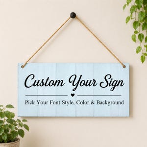 Könnte beinhalten: Ein rechteckiges, hellblaues Holzschild mit vertikaler Bretteroptik, das an einem beigen Seil hängt. Der Text "Custom Your Sign" steht in schwarzer Schrift über einem kleinen Herz, darunter "Pick Your Font Style, Color & Background".