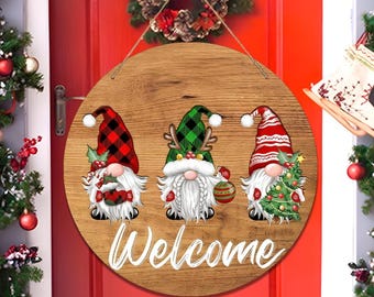 May include: A round wooden sign with three gnome figures wearing plaid hats and holding holiday decorations. The word "Welcome" is written in white script below the gnomes. The sign is hanging on a red door.