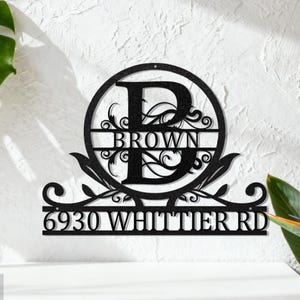 May include: A black metal house sign featuring the letter "D" within a circular design, with the name "BROWN" and address "6930 WHITTIER RD" below. The sign has decorative elements and is displayed against a textured white wall.