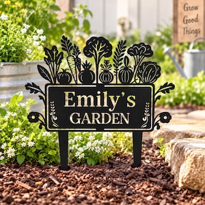 Puede incluir: Un estaca de jardín de metal negro con un letrero rectangular que muestra el texto "Emily's GARDEN" en una fuente serif. La parte superior del letrero está decorada con un diseño de silueta de varias verduras, incluyendo zanahorias y tomates.