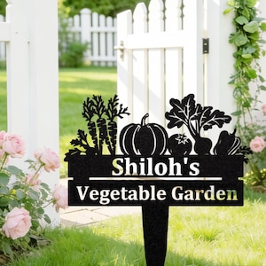Puede incluir: Letrero de jardín de metal negro con el texto "Shiloh's Vegetable Garden". El letrero presenta siluetas de verduras: zanahorias, calabaza, tomate, remolacha y pimiento. El letrero está en un jardín, con una valla blanca al fondo.