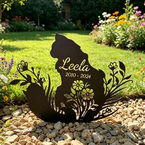 May include: A black metal cat silhouette garden stake with the name "Leela" and the years "2010-2024" in gold script. The cat is surrounded by floral and plant cutouts, set in a garden with flowers and grass.